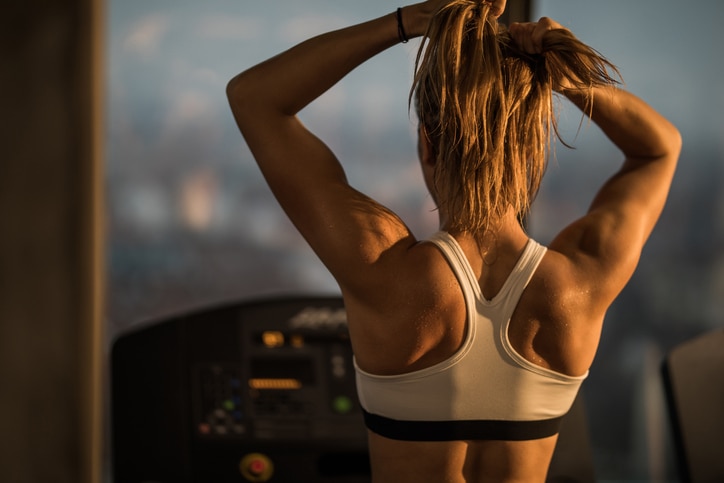 Rear view of muscular build woman tightening her ponytail before exercising on treadmill in a health club.