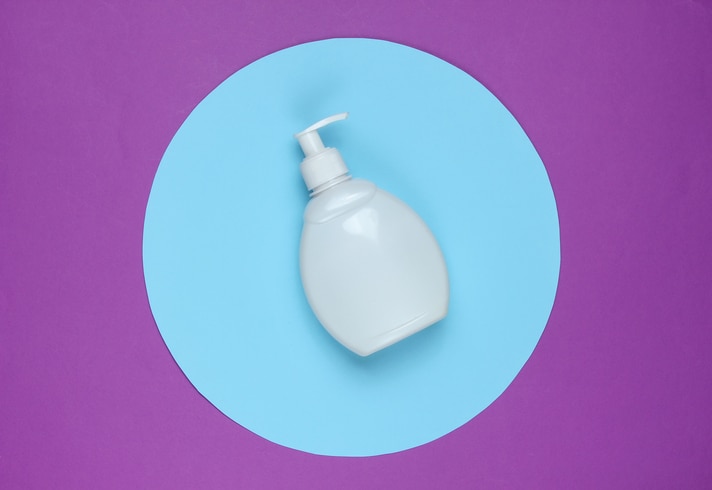 White shampoo bottle on a purple background with a blue pastel circle in the middle. Top view. Minimalistic beauty concept.