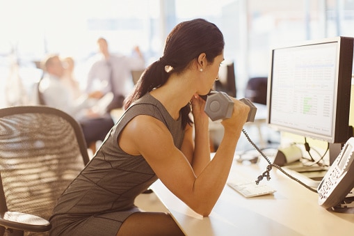 Businesswoman doing biceps curls with dumbbell and talking on telephone at computer in office