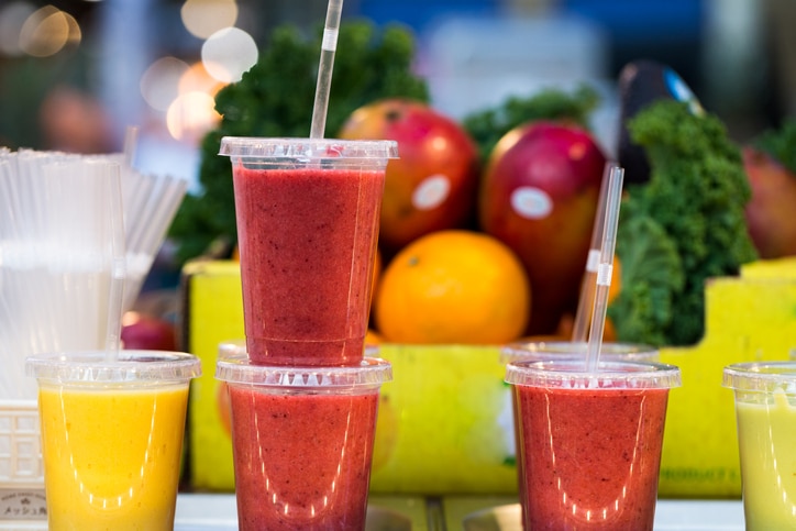 Close up color image depicting freshly made fruit juices and smoothies on display in a row and for sale at Spitalfields Market, a food and vintage market in east London, UK. In the background, defocused, is a pile of fresh fruit and vegetables - the raw ingredients used to make the smoothies. Room for copy space.