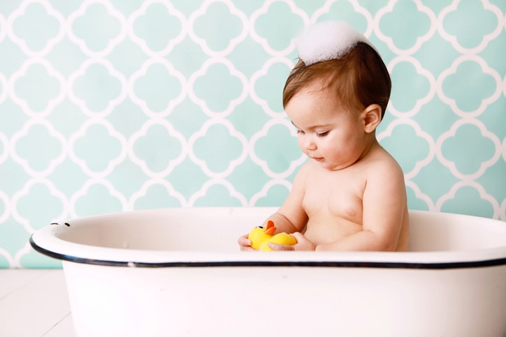 A baby looks at a rubber ducky as she sits in an antique bathtub. Copy space in vintage wallpaper.