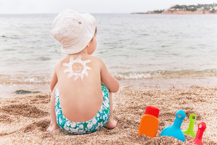 The sun drawing sunscreen (suntan lotion) on baby (boy)  back. Caucasian child is sitting with plastic container of sunscreen and toys on sunny beach. Close up, outdoor (Sharm El Sheikh, Egypt).