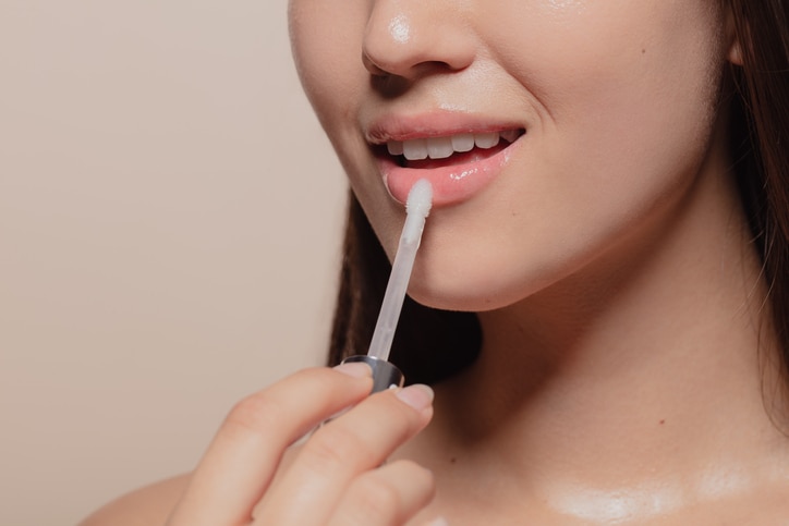 Close up of young korean woman applying transparent lip gloss. Cropped shot of girl putting on makeup on her lips with applicator against beige background.