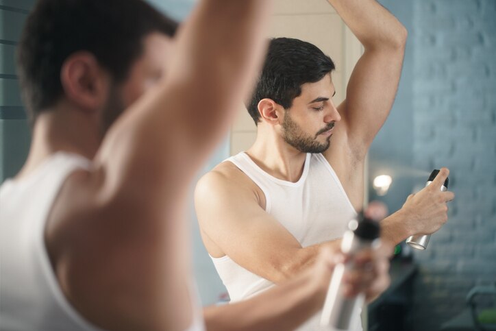 Young hispanic people and male beauty. Confident metrosexual man using spray deodorant on underarm skin, smiling and looking at mirror.