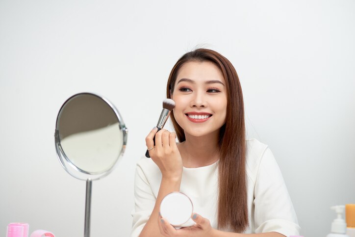 Beauty woman applying makeup. Beautiful girl looking in the mirror and applying cosmetic with a big brush. Girl gets blush on the cheekbones. Powder, rouge