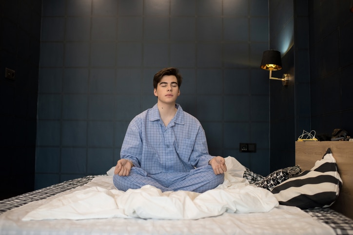 Portrait of teenage boy wearing pajamas meditating with eyes closed while sitting on bed in lotus position trying to sleep at night