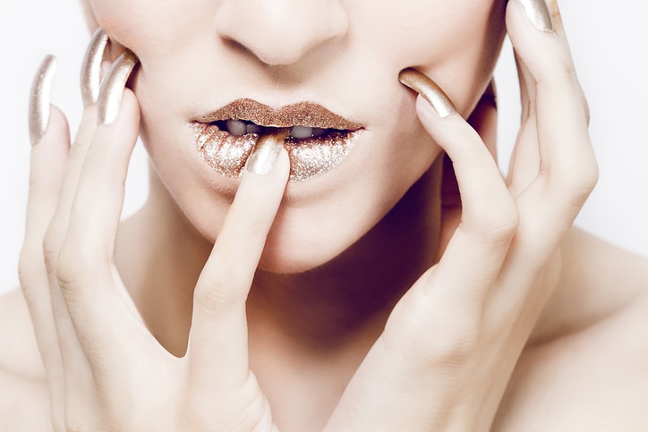 Closeup of beautiful woman with golden glitter lips and golden metallic nails, biting into nail.