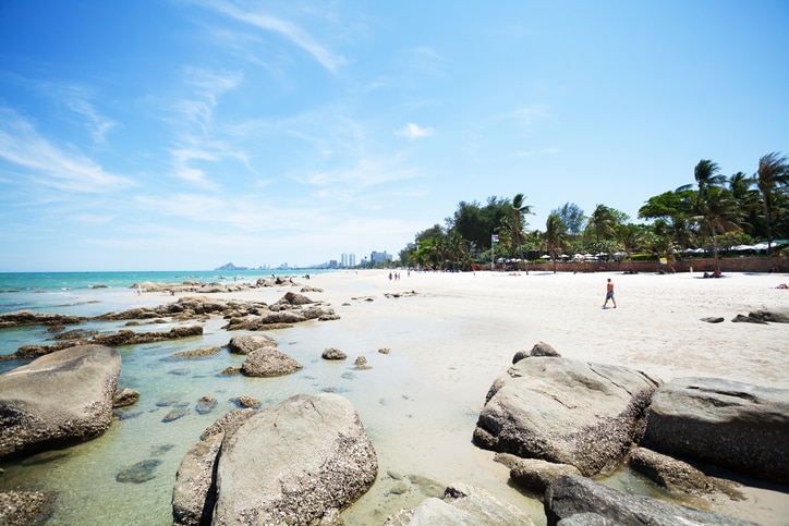 Hua Hin, Thailand - April 3, 2015: Capture of people on Hua Hin beach at sunny day. In foreground are rocks in sand and sea. View along beach to south. At right side are palm trees and resorts. In far background are hotels along coast.