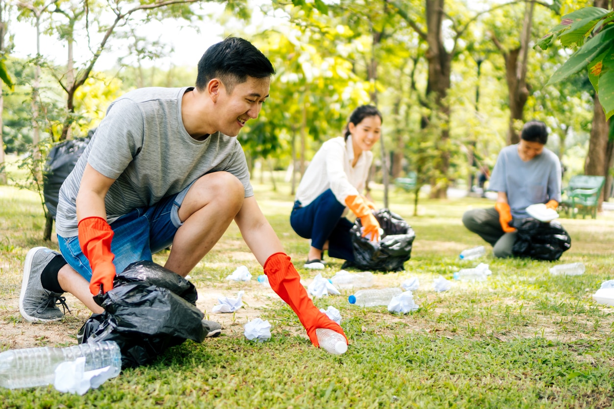 Young Asian man and women wearing orange gloves and collecting trash in garbage bag in the park. volunteer